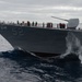 A U.S. Navy warship bearing hull number 52 is underway at sea, cutting through dark blue ocean waters with visible bow waves and spray. Multiple crew members in colored safety gear are visible on the deck, and a naval gun turret is mounted on the bow. The overcast sky and choppy seas suggest open ocean conditions.