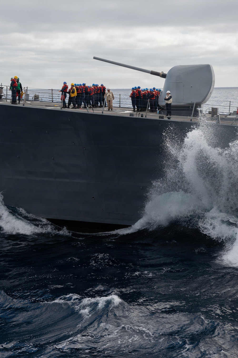 A naval warship is underway at sea in rough conditions, with its bow cutting through large waves and generating significant spray. Numerous crew members are assembled on the forward deck wearing orange life vests and blue hard hats, with a few supervisory personnel in white and yellow hard hats. A large naval gun turret (appears to be a 5-inch/127mm deck gun) is prominently visible at the bow.