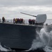 A naval warship is underway at sea in rough conditions, with its bow cutting through large waves and generating significant spray. Numerous crew members are assembled on the forward deck wearing orange life vests and blue hard hats, with a few supervisory personnel in white and yellow hard hats. A large naval gun turret (appears to be a 5-inch/127mm deck gun) is prominently visible at the bow.