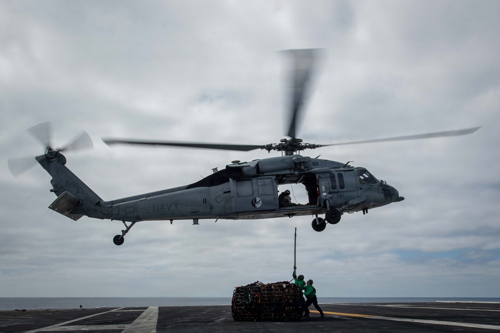 A U.S. Navy helicopter, marked 'NAVY' with tail number 611, hovers low over a ship's flight deck while conducting a vertical replenishment (VERTREP) operation. Two deck crew members in green jerseys are managing a cargo net loaded with supplies suspended beneath the aircraft. The overcast sky and open ocean horizon suggest an at-sea underway replenishment operation.