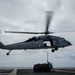 A U.S. Navy helicopter, marked 'NAVY' with tail number 611, hovers low over a ship's flight deck while conducting a vertical replenishment (VERTREP) operation. Two deck crew members in green jerseys are managing a cargo net loaded with supplies suspended beneath the aircraft. The overcast sky and open ocean horizon suggest an at-sea underway replenishment operation.
