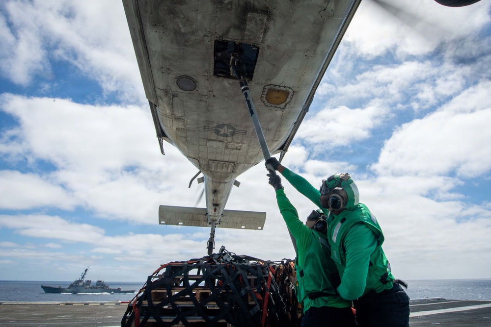 Two naval personnel in green jerseys and protective gear are working beneath the fuselage of a military aircraft on what appears to be an aircraft carrier flight deck. One worker is using a long pole to interact with a panel or component on the underside of the aircraft, while cargo in a net is visible nearby. A naval destroyer or escort vessel is visible in the background on open ocean.