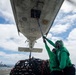 Two naval personnel in green jerseys and protective gear are working beneath the fuselage of a military aircraft on what appears to be an aircraft carrier flight deck. One worker is using a long pole to interact with a panel or component on the underside of the aircraft, while cargo in a net is visible nearby. A naval destroyer or escort vessel is visible in the background on open ocean.