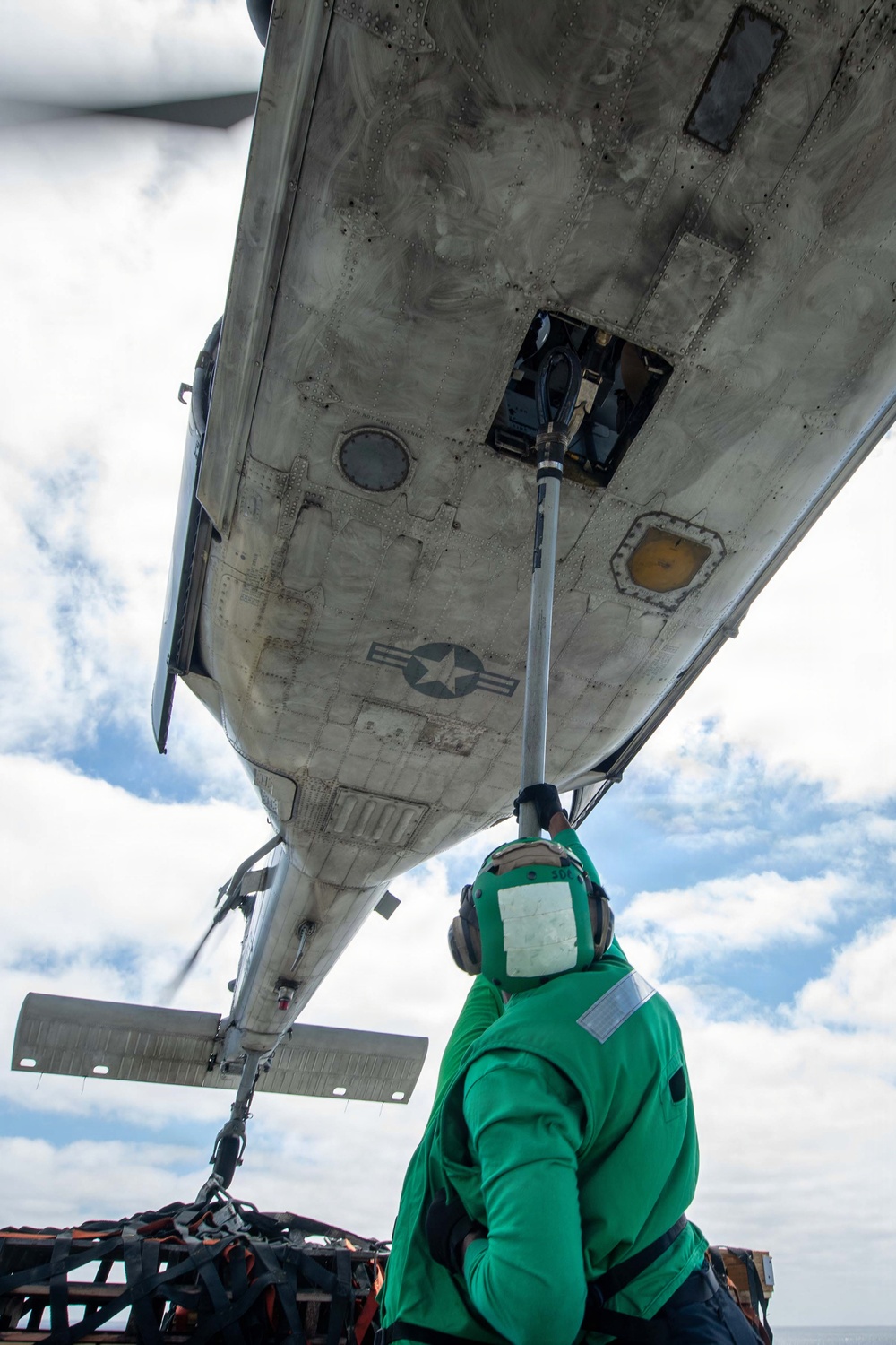 A ground crew member wearing a green jersey and hearing protection is using a long pole tool to service the underside of a military aircraft, visible from a low-angle upward perspective. The US military roundel (star and bar insignia) is clearly visible on the aircraft's fuselage underside. The setting appears to be an aircraft carrier flight deck, suggested by the tie-down chains and netting visible at the bottom of the frame.