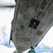 A ground crew member wearing a green jersey and hearing protection is using a long pole tool to service the underside of a military aircraft, visible from a low-angle upward perspective. The US military roundel (star and bar insignia) is clearly visible on the aircraft's fuselage underside. The setting appears to be an aircraft carrier flight deck, suggested by the tie-down chains and netting visible at the bottom of the frame.