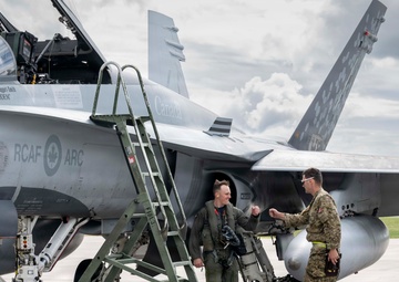 Two military personnel are standing beside a Royal Canadian Air Force (RCAF) CF-18 Hornet fighter jet on a tarmac. One individual appears to be a pilot in a flight suit carrying a helmet, while the other is in camouflage fatigues, and they appear to be engaged in a fist-bump or handshake gesture. The aircraft bears visible RCAF markings, the Canadian wordmark, and a tail number ending in '94'.
