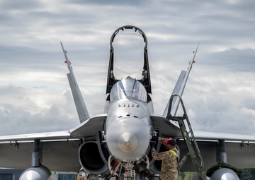 A military jet fighter, consistent with an F/A-18 Hornet or Super Hornet, is parked on an airfield tarmac with its canopy open. A ground crew technician in camouflage uniform is performing maintenance or pre-flight checks on the aircraft's nose/cockpit area using a maintenance ladder. The image is taken from a direct frontal perspective, providing a clear view of the aircraft's twin-engine intake configuration, wing-mounted fuel tanks, and landing gear.