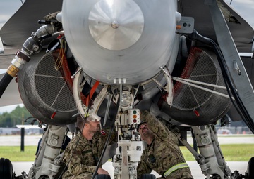Two military personnel in Navy Working Uniform (NWU) camouflage are conducting a maintenance inspection on the undercarriage of a military jet aircraft, likely an F/A-18 Hornet or Super Hornet. They are crouched beneath the fuselage examining the nose landing gear assembly and surrounding mechanical components. The aircraft is parked on a tarmac at what appears to be a military airfield.