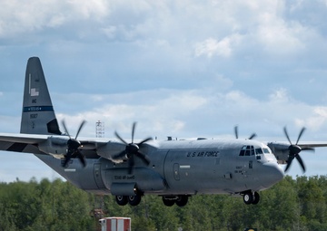 A U.S. Air Force C-130J Super Hercules military transport aircraft belonging to the Texas Air National Guard is captured during a landing or low-altitude approach on a runway. The aircraft is in a standard gray military paint scheme with markings clearly visible on the fuselage and tail. The setting appears to be a military or civilian airfield surrounded by dense tree lines under partly cloudy skies.