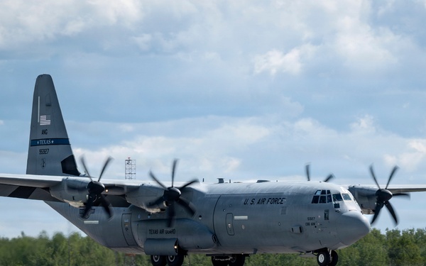 A U.S. Air Force C-130J Super Hercules military transport aircraft belonging to the Texas Air National Guard is captured during a landing or low-altitude approach on a runway. The aircraft is in a standard gray military paint scheme with markings clearly visible on the fuselage and tail. The setting appears to be a military or civilian airfield surrounded by dense tree lines under partly cloudy skies.