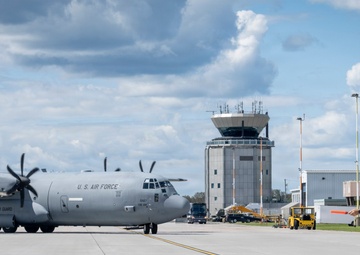 A U.S. Air Force C-130 Hercules transport aircraft is being marshaled on a military airfield tarmac by a ground crew member holding orange wands. The aircraft bears 'U.S. AIR FORCE' and 'TEXAS AIR GUARD' markings, indicating it belongs to the Texas Air National Guard. A control tower and base operations buildings are visible in the background.