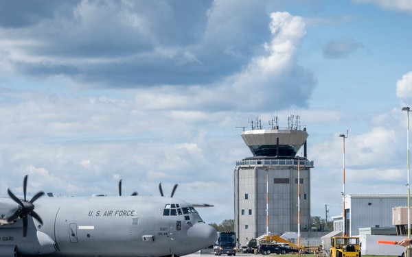 A U.S. Air Force C-130 Hercules transport aircraft is being marshaled on a military airfield tarmac by a ground crew member holding orange wands. The aircraft bears 'U.S. AIR FORCE' and 'TEXAS AIR GUARD' markings, indicating it belongs to the Texas Air National Guard. A control tower and base operations buildings are visible in the background.
