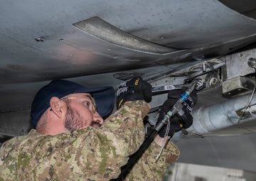 A military technician in camouflage uniform is performing maintenance work on the underside of an aircraft inside a hangar. The individual is using a pneumatic or hydraulic tool to work on components mounted beneath the aircraft's fuselage or wing. The setting appears to be an indoor maintenance facility with equipment visible in the background.