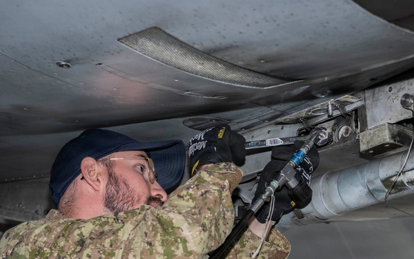 A military technician in camouflage uniform is performing maintenance work on the underside of an aircraft inside a hangar. The individual is using a pneumatic or hydraulic tool to work on components mounted beneath the aircraft's fuselage or wing. The setting appears to be an indoor maintenance facility with equipment visible in the background.