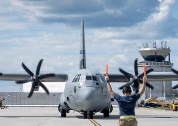A military ground crew member is marshaling a large turboprop military transport aircraft, identifiable as a C-130J Super Hercules, on a tarmac. The marshal is using orange signal wands with both arms raised, guiding the aircraft toward its parking position. A control tower, ground support equipment, and a red helicopter are visible in the background.
