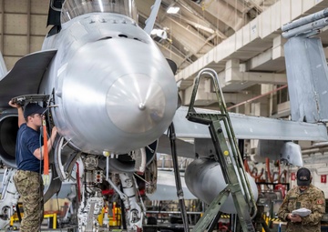 Two military personnel are performing maintenance on a fighter jet inside a large hangar facility. One technician is standing on a blue step ladder working on the nose/avionics bay area of the aircraft, while a second person reviews documentation nearby. The aircraft appears to be an F/A-18 Hornet or Super Hornet, identifiable by its distinctive twin-engine nose configuration and gray paint scheme.