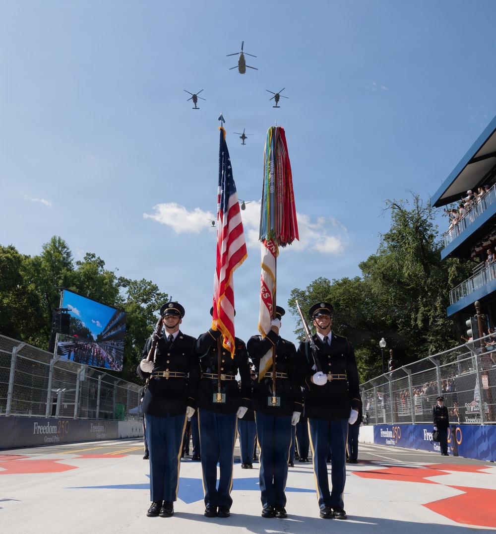 A military color guard ceremony is taking place at the 'Freedom 250 Grand Prix' racing event, with uniformed soldiers presenting the American flag and a military unit colors flag. A formation of five helicopters is conducting a flyover directly above the color guard in a V-shaped formation. The event appears to be held on a racing circuit with grandstands filled with spectators visible in the background.