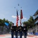 A military color guard ceremony is taking place at the 'Freedom 250 Grand Prix' racing event, with uniformed soldiers presenting the American flag and a military unit colors flag. A formation of five helicopters is conducting a flyover directly above the color guard in a V-shaped formation. The event appears to be held on a racing circuit with grandstands filled with spectators visible in the background.