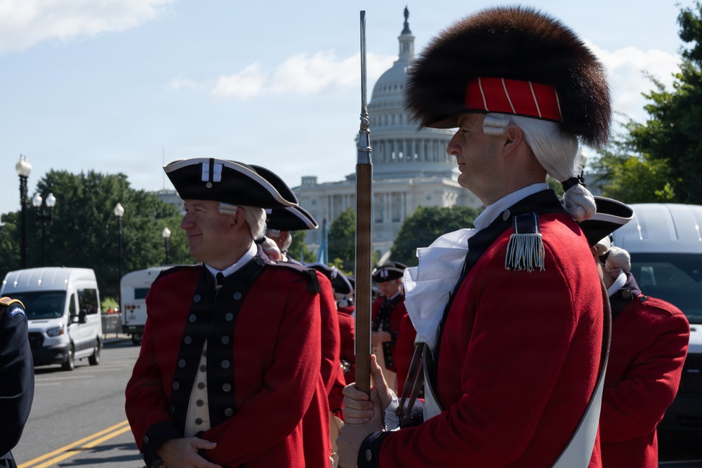 Two men dressed in 18th-century American Revolutionary War-era military uniforms stand on a street in Washington, D.C., with the U.S. Capitol building visible in the background. The foreground figure holds a period-accurate musket and wears a distinctive fur-topped grenadier cap, while the other wears a tricorn hat. This appears to be a ceremonial parade or reenactment event near the Capitol.