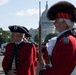 Two men dressed in 18th-century American Revolutionary War-era military uniforms stand on a street in Washington, D.C., with the U.S. Capitol building visible in the background. The foreground figure holds a period-accurate musket and wears a distinctive fur-topped grenadier cap, while the other wears a tricorn hat. This appears to be a ceremonial parade or reenactment event near the Capitol.