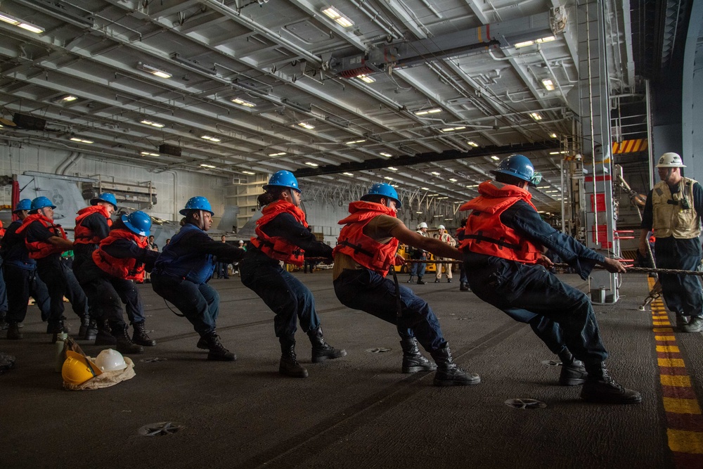 A group of naval personnel are performing a line-pulling drill inside what appears to be the hangar bay of a large warship, likely an aircraft carrier. The sailors are wearing orange life vests and blue hard hats while hauling on a thick rope in a coordinated team effort. A supervisor in a white hard hat observes from the right side of the frame.