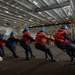 A group of naval personnel are performing a line-pulling drill inside what appears to be the hangar bay of a large warship, likely an aircraft carrier. The sailors are wearing orange life vests and blue hard hats while hauling on a thick rope in a coordinated team effort. A supervisor in a white hard hat observes from the right side of the frame.