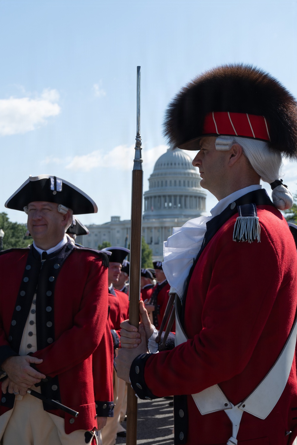 A group of individuals dressed in 18th-century American Revolutionary War-era military reenactment uniforms are gathered outdoors in front of the United States Capitol building in Washington, D.C. The participants wear red coats, tricorn hats, and bearskin caps, and carry period-accurate flintlock muskets with bayonets. The scene appears to be a ceremonial event or historical reenactment on a clear, sunny day.