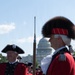 A group of individuals dressed in 18th-century American Revolutionary War-era military reenactment uniforms are gathered outdoors in front of the United States Capitol building in Washington, D.C. The participants wear red coats, tricorn hats, and bearskin caps, and carry period-accurate flintlock muskets with bayonets. The scene appears to be a ceremonial event or historical reenactment on a clear, sunny day.