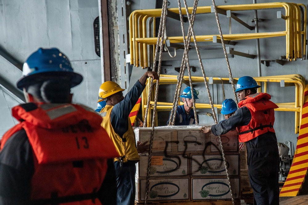 Military or naval personnel are conducting a replenishment-at-sea (RAS) or cargo transfer operation aboard a ship. Workers wearing hard hats and orange life vests are handling a pallet of boxed goods suspended by rope netting. The boxes are visibly labeled 'Mayor's Choice Lettuce' from California, indicating a food supply transfer.