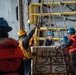 Military or naval personnel are conducting a replenishment-at-sea (RAS) or cargo transfer operation aboard a ship. Workers wearing hard hats and orange life vests are handling a pallet of boxed goods suspended by rope netting. The boxes are visibly labeled 'Mayor's Choice Lettuce' from California, indicating a food supply transfer.