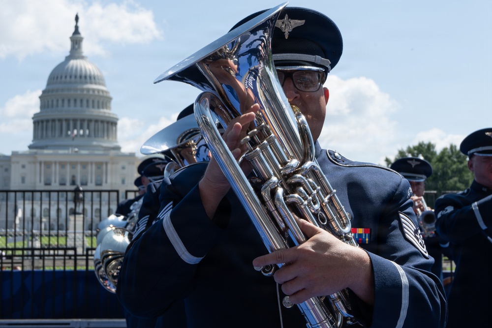 A military band performs outdoors in front of the United States Capitol building in Washington, D.C. The foreground musician is playing a euphonium or baritone horn while wearing a dark blue dress uniform with service ribbons and a peaked cap bearing a military insignia. Multiple band members are visible in formation behind the lead musician.