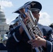 A military band performs outdoors in front of the United States Capitol building in Washington, D.C. The foreground musician is playing a euphonium or baritone horn while wearing a dark blue dress uniform with service ribbons and a peaked cap bearing a military insignia. Multiple band members are visible in formation behind the lead musician.