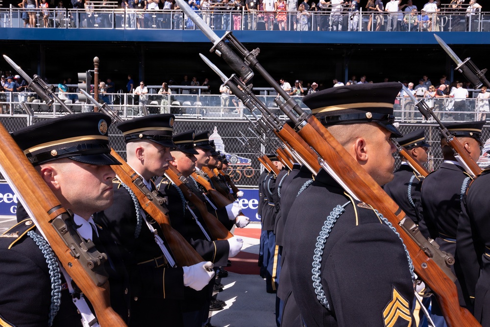 A military honor guard unit is performing at a formal ceremonial parade or event, with soldiers dressed in dark dress uniforms carrying rifles with fixed bayonets. The soldiers are wearing distinctive service caps with gold trim and blue aiguillettes, suggesting this is a U.S. Army ceremonial unit. A large crowd of spectators is visible in tiered bleachers in the background.