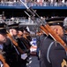 A military honor guard unit is performing at a formal ceremonial parade or event, with soldiers dressed in dark dress uniforms carrying rifles with fixed bayonets. The soldiers are wearing distinctive service caps with gold trim and blue aiguillettes, suggesting this is a U.S. Army ceremonial unit. A large crowd of spectators is visible in tiered bleachers in the background.