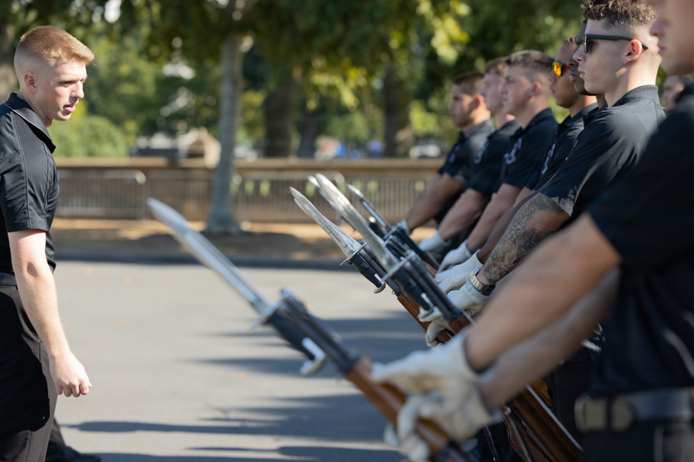 A group of uniformed individuals in black shirts are standing in a formal line holding rifles with bayonets attached, while a young man faces them in what appears to be a drill or inspection exercise. The participants are wearing white gloves, suggesting a ceremonial or honor guard context. The setting appears to be an outdoor paved area with trees visible in the background.