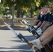 A group of uniformed individuals in black shirts are standing in a formal line holding rifles with bayonets attached, while a young man faces them in what appears to be a drill or inspection exercise. The participants are wearing white gloves, suggesting a ceremonial or honor guard context. The setting appears to be an outdoor paved area with trees visible in the background.