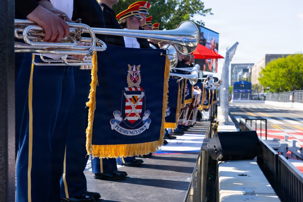 A military band identified as 'Pershing's Own' — the U.S. Army Band — is lined up on an outdoor stage, holding silver herald trumpets adorned with navy blue and gold-fringed banners bearing the unit's coat of arms. The musicians are dressed in formal military uniforms with blue trousers featuring gold stripes, and some wear red-plumed ceremonial hats. The setting appears to be a public outdoor ceremony or parade event in an urban environment.