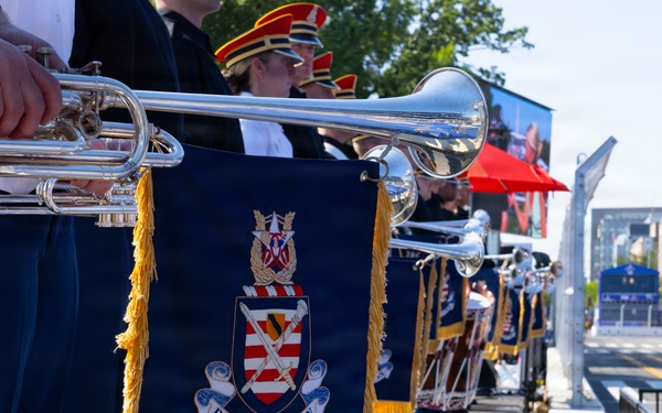 A military band identified as 'Pershing's Own' — the U.S. Army Band — is lined up on an outdoor stage, holding silver herald trumpets adorned with navy blue and gold-fringed banners bearing the unit's coat of arms. The musicians are dressed in formal military uniforms with blue trousers featuring gold stripes, and some wear red-plumed ceremonial hats. The setting appears to be a public outdoor ceremony or parade event in an urban environment.