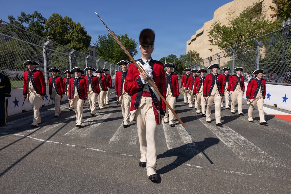 A precision drill unit dressed in 18th-century Continental Army-style uniforms — red coats, cream breeches, tricorn hats, and tall bearskin caps — marches in formation along a paved surface. The lead soldier carries a long ceremonial musket or pike and wears a distinctive bearskin cap, suggesting a drum major or color guard role. The setting appears to be an outdoor ceremonial parade or public event, likely in Washington, D.C., based on the visible architecture.