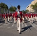 A precision drill unit dressed in 18th-century Continental Army-style uniforms — red coats, cream breeches, tricorn hats, and tall bearskin caps — marches in formation along a paved surface. The lead soldier carries a long ceremonial musket or pike and wears a distinctive bearskin cap, suggesting a drum major or color guard role. The setting appears to be an outdoor ceremonial parade or public event, likely in Washington, D.C., based on the visible architecture.