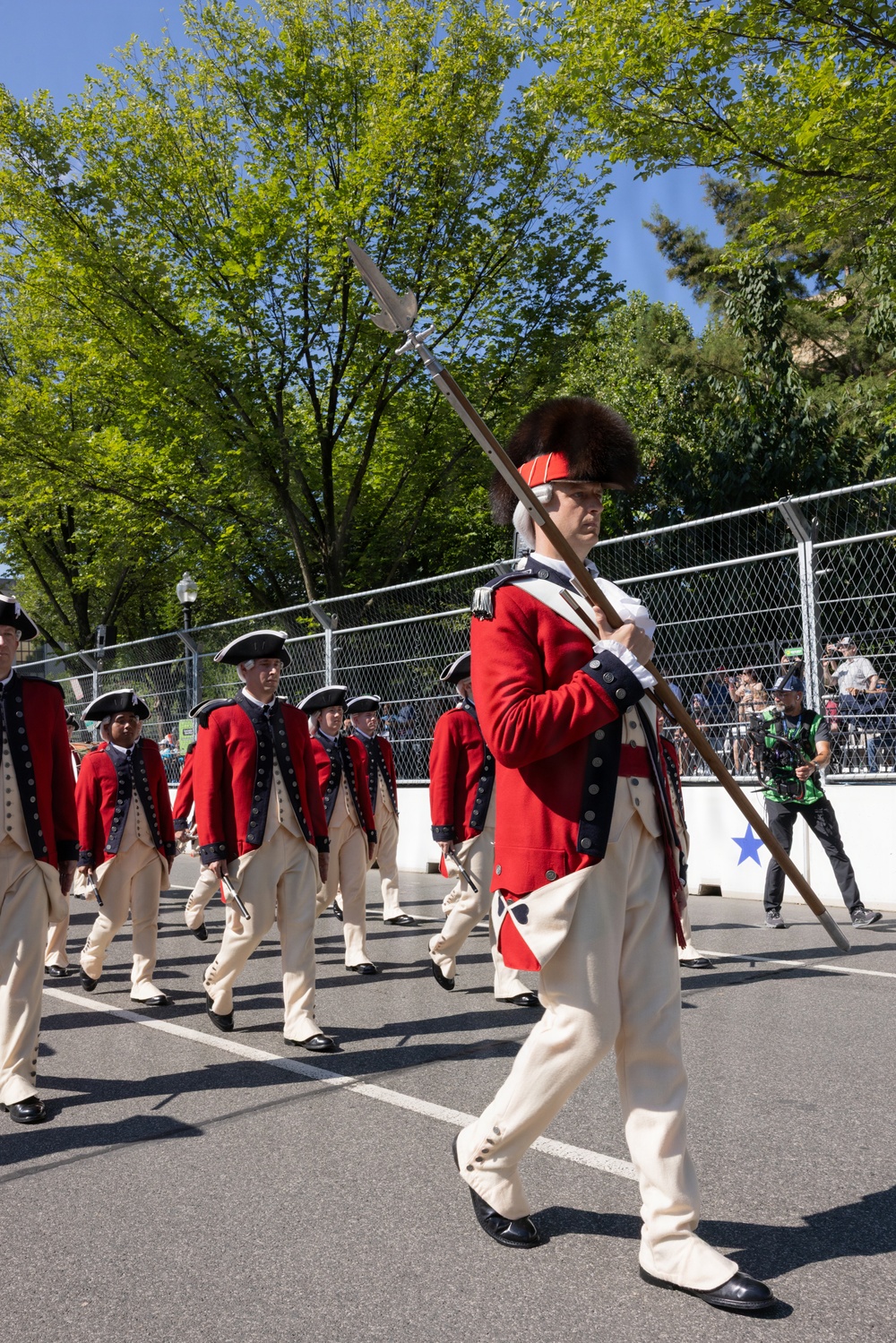 A group of performers dressed in 18th-century American Revolutionary War-era military uniforms march in a parade along a paved street. The lead figure wears a distinctive tall bearskin cap and carries a ceremonial halberd/spear, while the others wear tricorn hats and carry rifles or drumsticks. The setting appears to be a modern outdoor public event with spectators, chain-link fencing, and trees visible in the background.