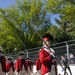 A group of performers dressed in 18th-century American Revolutionary War-era military uniforms march in a parade along a paved street. The lead figure wears a distinctive tall bearskin cap and carries a ceremonial halberd/spear, while the others wear tricorn hats and carry rifles or drumsticks. The setting appears to be a modern outdoor public event with spectators, chain-link fencing, and trees visible in the background.