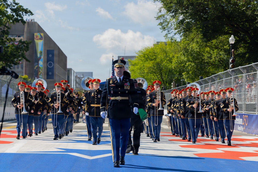 A U.S. Army military marching band performs in a formal parade along a decorated street with red, white, and blue markings. The band members wear distinctive dark uniforms with red-plumed caps, led by a decorated officer in full dress uniform with white gloves. A banner visible in the background reads 'Freedom 250,' suggesting this is a commemorative patriotic event.