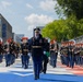 A U.S. Army military marching band performs in a formal parade along a decorated street with red, white, and blue markings. The band members wear distinctive dark uniforms with red-plumed caps, led by a decorated officer in full dress uniform with white gloves. A banner visible in the background reads 'Freedom 250,' suggesting this is a commemorative patriotic event.