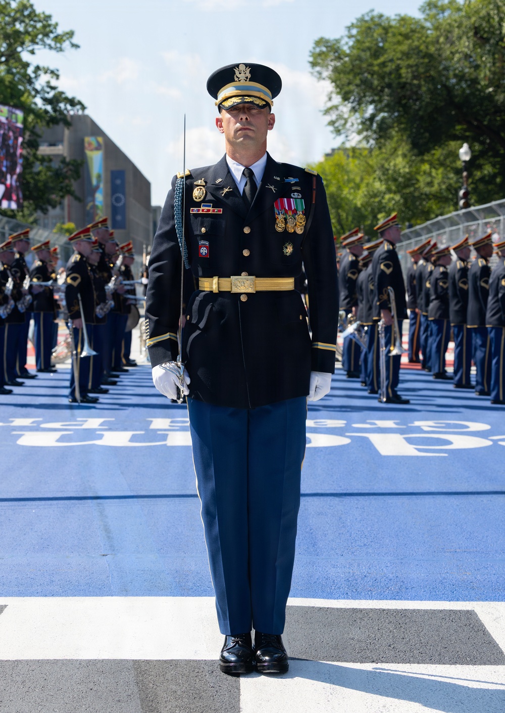 A U.S. Army officer in full dress blues uniform stands at attention on a blue-painted street surface, holding a ceremonial saber. He is positioned at the front of a military formation during what appears to be a formal parade or ceremonial event in an urban setting. Rows of soldiers in matching dress uniforms are visible on both sides behind him.