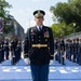 A U.S. Army officer in full dress blues uniform stands at attention on a blue-painted street surface, holding a ceremonial saber. He is positioned at the front of a military formation during what appears to be a formal parade or ceremonial event in an urban setting. Rows of soldiers in matching dress uniforms are visible on both sides behind him.
