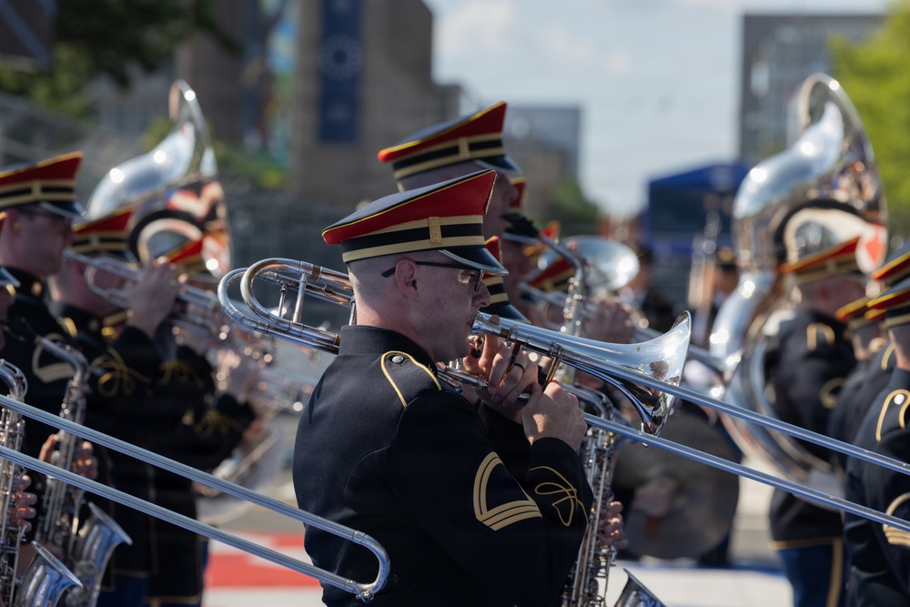 A military marching band is performing outdoors, likely during a parade or public ceremony in an urban setting. Musicians are dressed in formal black uniforms with red and gold-trimmed shakos (caps) and are playing brass instruments. The central figure in focus is playing a flugelhorn or trumpet while trombone slides are visible in the foreground.