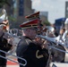 A military marching band is performing outdoors, likely during a parade or public ceremony in an urban setting. Musicians are dressed in formal black uniforms with red and gold-trimmed shakos (caps) and are playing brass instruments. The central figure in focus is playing a flugelhorn or trumpet while trombone slides are visible in the foreground.