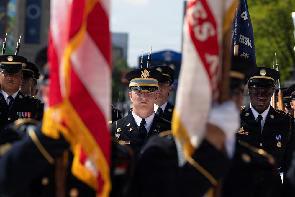 A formal U.S. Army color guard or honor guard unit is participating in a military parade or ceremonial event. Soldiers in dress black uniforms with service caps are marching in formation, carrying flags including what appears to be the U.S. flag and unit guidons. The central figure, an officer identifiable by his gold-banded cap and officer insignia, leads the formation with a composed, forward-focused expression.