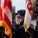 A formal U.S. Army color guard or honor guard unit is participating in a military parade or ceremonial event. Soldiers in dress black uniforms with service caps are marching in formation, carrying flags including what appears to be the U.S. flag and unit guidons. The central figure, an officer identifiable by his gold-banded cap and officer insignia, leads the formation with a composed, forward-focused expression.